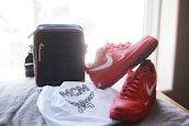 A pair of bright red Nike sneakers with white swooshes are positioned next to a black handbag with red trim. In front of these items is a white dust bag with a logo and the letters 'MCM' printed on it. The items are set against a soft, beige surface near a window with soft natural light streaming in.