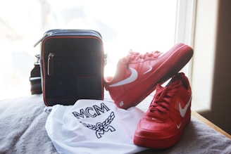A pair of bright red Nike sneakers with white swooshes are positioned next to a black handbag with red trim. In front of these items is a white dust bag with a logo and the letters 'MCM' printed on it. The items are set against a soft, beige surface near a window with soft natural light streaming in.