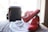 A pair of bright red Nike sneakers with white swooshes are positioned next to a black handbag with red trim. In front of these items is a white dust bag with a logo and the letters 'MCM' printed on it. The items are set against a soft, beige surface near a window with soft natural light streaming in.