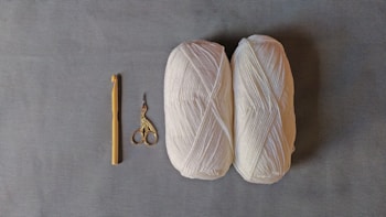 Two skeins of white yarn are placed on a gray surface alongside a yellow crochet hook and a pair of ornate scissors with a bird design.