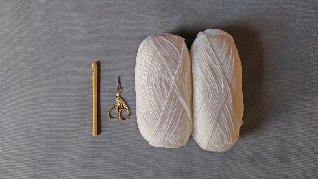 Two skeins of white yarn are placed on a gray surface alongside a yellow crochet hook and a pair of ornate scissors with a bird design.