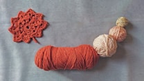 A collection of yarn balls in various sizes, featuring colors such as red, pink, and beige, arranged in a line on a plain surface. To the left, there is a red crocheted pattern resembling a flower.