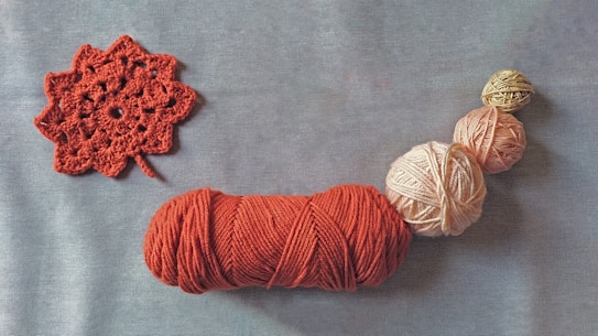 A collection of yarn balls in various sizes, featuring colors such as red, pink, and beige, arranged in a line on a plain surface. To the left, there is a red crocheted pattern resembling a flower.