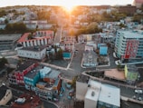 Aerial view of a colorful urban neighborhood with various buildings painted in red, blue, yellow, and teal. Streets intersect in a grid pattern with a few cars visible. Lush green trees are interspersed among the buildings, and the sun sets on the horizon, casting a warm glow over the scene.