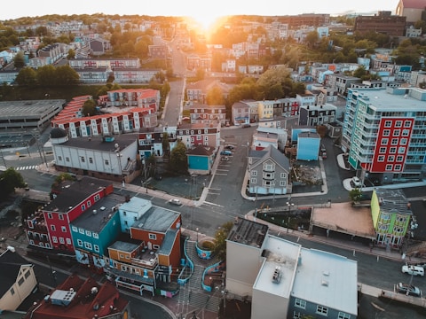 Aerial view of a colorful urban neighborhood with various buildings painted in red, blue, yellow, and teal. Streets intersect in a grid pattern with a few cars visible. Lush green trees are interspersed among the buildings, and the sun sets on the horizon, casting a warm glow over the scene.