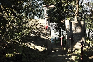 A narrow pathway flanked by tall concrete walls leads through a wooded area. Sunlight filters through the dense foliage, casting intricate shadows on the walls and ground. A light fixture is mounted above the path, and a red fire extinguisher is attached to one of the walls. In the distance, light hints at an open area.