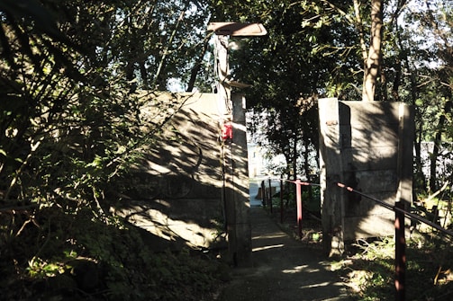 A narrow pathway flanked by tall concrete walls leads through a wooded area. Sunlight filters through the dense foliage, casting intricate shadows on the walls and ground. A light fixture is mounted above the path, and a red fire extinguisher is attached to one of the walls. In the distance, light hints at an open area.