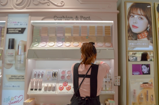 A woman is standing in front of a cosmetics display in a store, selecting a product. The shelves are filled with various foundation compacts, powders, and other beauty products. Advertisements and promotional images featuring models are visible on the surrounding walls.