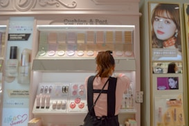 A woman is standing in front of a cosmetics display in a store, selecting a product. The shelves are filled with various foundation compacts, powders, and other beauty products. Advertisements and promotional images featuring models are visible on the surrounding walls.