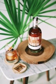 A bottle of Cask & Crew whiskey is placed on a wooden slab, accompanied by a glass filled with a whiskey cocktail garnished with cherries and an orange slice. The setup is on a small marble table, and a large green plant provides a lush background.