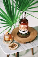 A bottle of Cask & Crew whiskey is placed on a wooden slab, accompanied by a glass filled with a whiskey cocktail garnished with cherries and an orange slice. The setup is on a small marble table, and a large green plant provides a lush background.