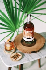 A bottle of Cask & Crew whiskey is placed on a wooden slab, accompanied by a glass filled with a whiskey cocktail garnished with cherries and an orange slice. The setup is on a small marble table, and a large green plant provides a lush background.