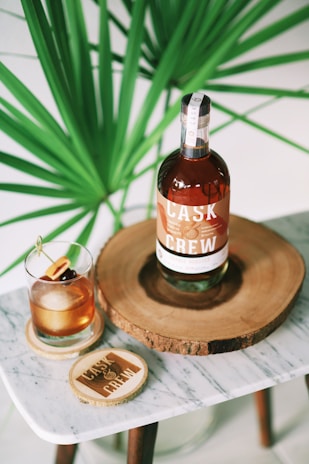 A bottle of Cask & Crew whiskey is placed on a wooden slab, accompanied by a glass filled with a whiskey cocktail garnished with cherries and an orange slice. The setup is on a small marble table, and a large green plant provides a lush background.