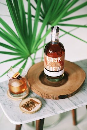 A bottle of Cask & Crew whiskey is placed on a wooden slab, accompanied by a glass filled with a whiskey cocktail garnished with cherries and an orange slice. The setup is on a small marble table, and a large green plant provides a lush background.