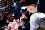A group of elderly men are seated around a wooden table outdoors, engaged in playing a card game. They appear to be focused and are holding cards in their hands. The table is covered with a cloth, and there are some discarded cards on the surface. The scene is well-lit by natural sunlight, creating a warm ambiance.
