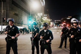 A group of police officers in uniform stand in formation on a city street at night. They are wearing helmets and holding batons. In the background, there is a building with bright lights and several people gathered near a traffic light.