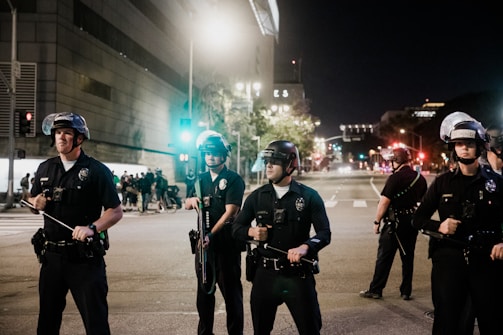 A group of police officers in uniform stand in formation on a city street at night. They are wearing helmets and holding batons. In the background, there is a building with bright lights and several people gathered near a traffic light.