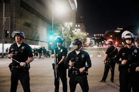 A group of police officers in uniform stand in formation on a city street at night. They are wearing helmets and holding batons. In the background, there is a building with bright lights and several people gathered near a traffic light.