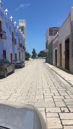 A cobblestone street flanked by colorful colonial buildings stretches towards a distant tree line. The street is lined with parked cars and illuminated by bright daylight. The buildings feature pastel colors such as blue, pink, and green, with decorative details and balconies.