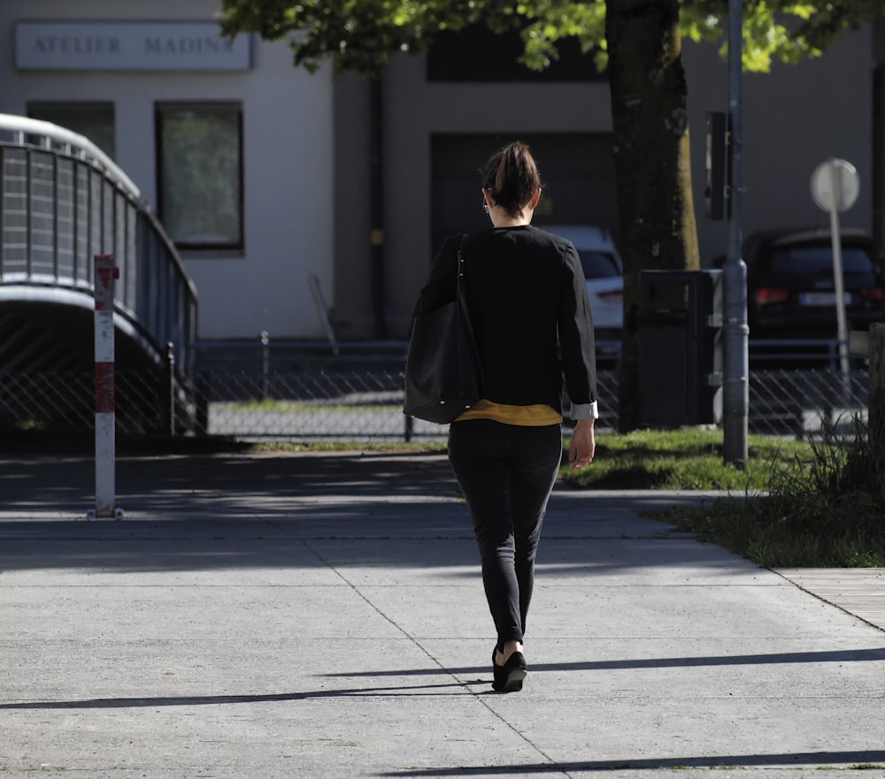 Mujer en chaqueta negra y pantalones negros caminando en la acera durante el día