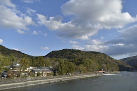 A serene landscape featuring a river flowing alongside a small village nestled at the base of verdant hills. The sky is partly cloudy, with sunlight casting soft shadows over the scene. The hills are lush with green vegetation, and the village consists of a few traditional-style buildings.