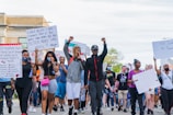 A diverse group of people is marching, holding various protest signs that call for justice and change. Many demonstrators have raised fists, indicating solidarity and determination. The participants are dressed casually and some wear masks. The setting appears to be an urban street.