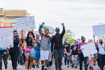 A diverse group of people is marching, holding various protest signs that call for justice and change. Many demonstrators have raised fists, indicating solidarity and determination. The participants are dressed casually and some wear masks. The setting appears to be an urban street.