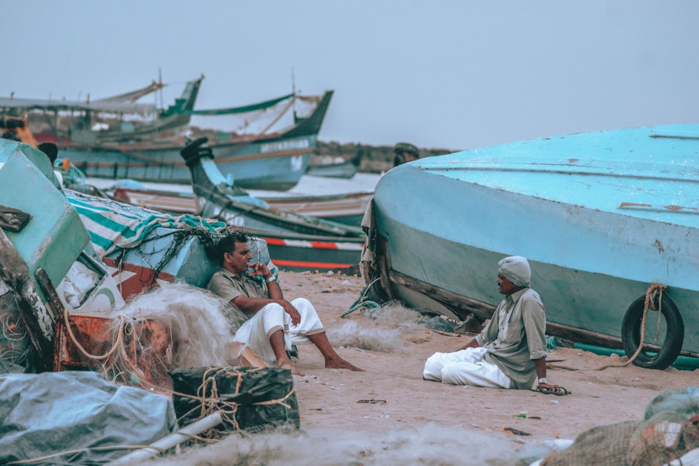 Mann in Grau Thobe sitzt tagsüber auf braunem Sand in der Nähe von Green Boat