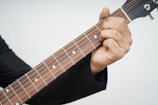 A hand wearing a black sleeve holds the neck of a guitar, pressing down on the strings at specific frets. The guitar's neck and strings are clearly visible, and the person appears to be forming a chord.