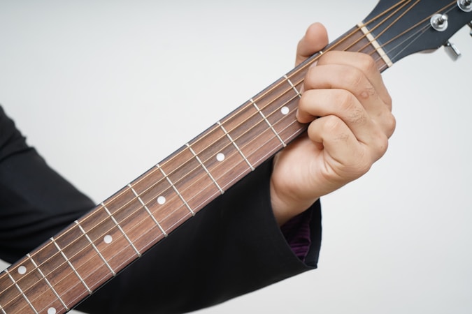 A hand wearing a black sleeve holds the neck of a guitar, pressing down on the strings at specific frets. The guitar's neck and strings are clearly visible, and the person appears to be forming a chord.