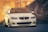 A sleek, modified white BMW car with customized features is parked on a smooth urban road. The car is highlighted against a blurred background of a brick wall, suggesting an urban environment. The vehicle's lowered stance and sport rims enhance its stylish, aggressive look.
