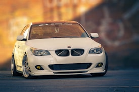 A sleek, modified white BMW car with customized features is parked on a smooth urban road. The car is highlighted against a blurred background of a brick wall, suggesting an urban environment. The vehicle's lowered stance and sport rims enhance its stylish, aggressive look.