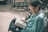 An elderly person with glasses is seated outdoors, engrossed in reading a book with well-thumbed pages. The background is slightly blurred, featuring wooden furniture and various objects.