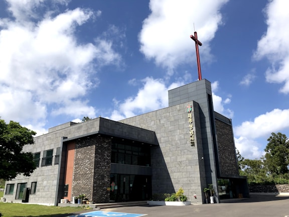 A modern church building with a gray stone facade and a tall red cross on the roof. It is a two-story structure with large glass windows and a landscaped area featuring small potted plants near the entrance. The sky is bright with fluffy white clouds.