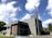 A modern church building with a gray stone facade and a tall red cross on the roof. It is a two-story structure with large glass windows and a landscaped area featuring small potted plants near the entrance. The sky is bright with fluffy white clouds.