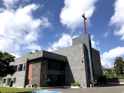 A modern church building with a gray stone facade and a tall red cross on the roof. It is a two-story structure with large glass windows and a landscaped area featuring small potted plants near the entrance. The sky is bright with fluffy white clouds.