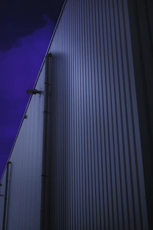 An evening 4K shot of a metal sheet shed with blue roofing and steel frame, with warm light glowing inside, located in an Indian industrial area.