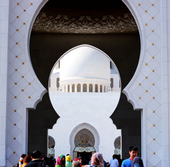An architectural design featuring an ornate archway with a view of a domed building in the background. The intricate patterns and geometric shapes highlight a traditional style. People are gathered at the base, adding a contrast to the grand architectural elements.