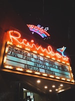A vintage-style theater marquee illuminated at night, featuring a neon sign with 'Rainbow' and a colorful rainbow design above it. The marquee displays announcements for events dated 16th of October, 2016, including names like Paul Paul Teatro Feria de Fresno, Intocable, and Lupillo Rivera. The scene has an atmospheric glow with warm yellow lights surrounding the sign.
