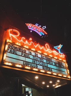 A vintage-style theater marquee illuminated at night, featuring a neon sign with 'Rainbow' and a colorful rainbow design above it. The marquee displays announcements for events dated 16th of October, 2016, including names like Paul Paul Teatro Feria de Fresno, Intocable, and Lupillo Rivera. The scene has an atmospheric glow with warm yellow lights surrounding the sign.