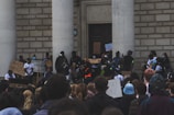 A group of people gathered outside a large building with two columns, holding signs with messages such as 'Black Lives Matter.' Many participants are wearing masks, and the gathering appears to be peaceful but serious. The crowd is diverse, and some people are using megaphones.