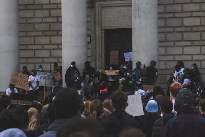 A group of people gathered outside a large building with two columns, holding signs with messages such as 'Black Lives Matter.' Many participants are wearing masks, and the gathering appears to be peaceful but serious. The crowd is diverse, and some people are using megaphones.