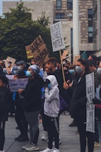 A diverse group of people gather outdoors, many wearing face masks. They hold signs with messages advocating for racial justice and social change, including 'Black Lives Matter' and 'Silence is Violence.' The setting is urban, with modern architecture and greenery in the background.