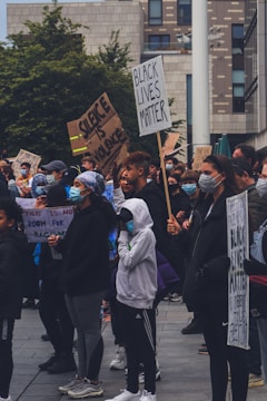 A diverse group of people gather outdoors, many wearing face masks. They hold signs with messages advocating for racial justice and social change, including 'Black Lives Matter' and 'Silence is Violence.' The setting is urban, with modern architecture and greenery in the background.