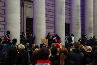 A group of people are gathered in front of a building with large columns. Several individuals are holding signs, and one sign reads 'Black Lives Matter.' Many in the crowd are wearing masks, and some appear to be giving speeches or holding a demonstration.