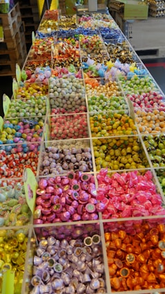 An arrangement of various colorful candies is displayed in clear compartments, creating a mosaic of vibrant hues. Each section is filled with neatly organized sweets, varying in size, shape, and color, including shiny wrappers and different textures.