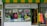 A storefront with bright yellow and green striped awning, featuring a Jamaican culture gift shop. The windows display various clothing items including T-shirts with Jamaican themes, mannequins dressed in vibrant colors, posters, and decorative items. A neon 'OPEN' sign is visible, alongside promotional signage for rum bottles and a sale.