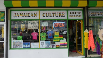 A storefront with bright yellow and green striped awning, featuring a Jamaican culture gift shop. The windows display various clothing items including T-shirts with Jamaican themes, mannequins dressed in vibrant colors, posters, and decorative items. A neon 'OPEN' sign is visible, alongside promotional signage for rum bottles and a sale.