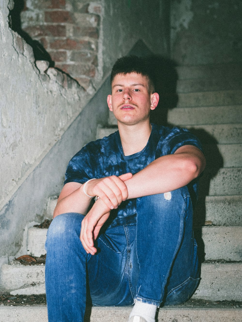 Hombre con camisa azul abotonada y jeans de mezclilla azul sentado en una pared de concreto