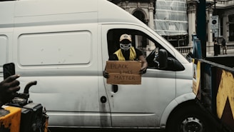 A person wearing a mask and neon vest is sitting in the driver's seat of a white van, holding a cardboard sign that reads 'Black Lives Matter'. Another person is partially visible, holding a smartphone and appearing to take a picture or video. The background features a building with classical architecture and some construction elements.